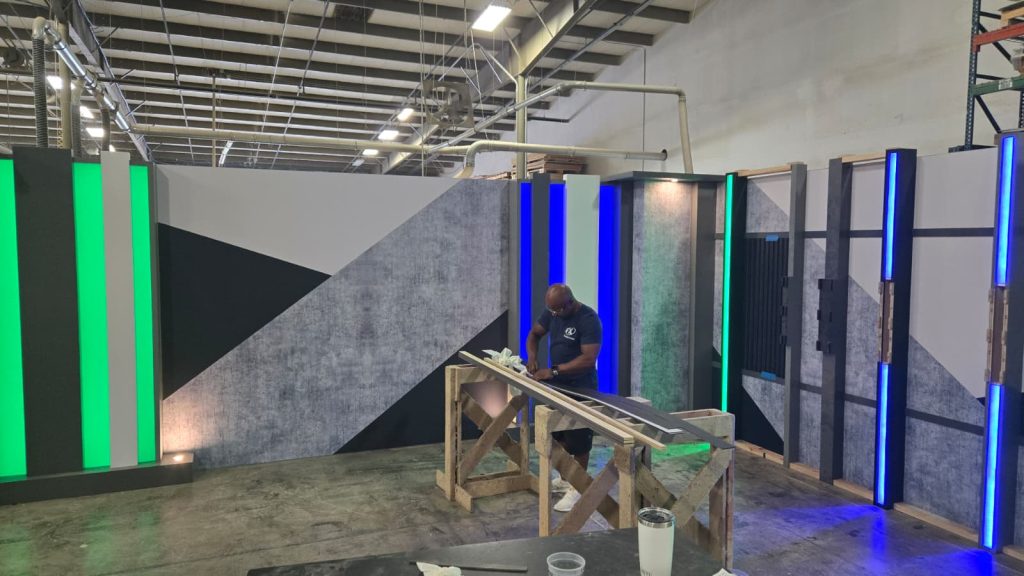 A worker meticulously assembling components of a contemporary broadcast studio set, featuring geometric wall panels with integrated green and blue LED lighting, highlighting FX Design Group's comprehensive set design, fabrication, and installation services.